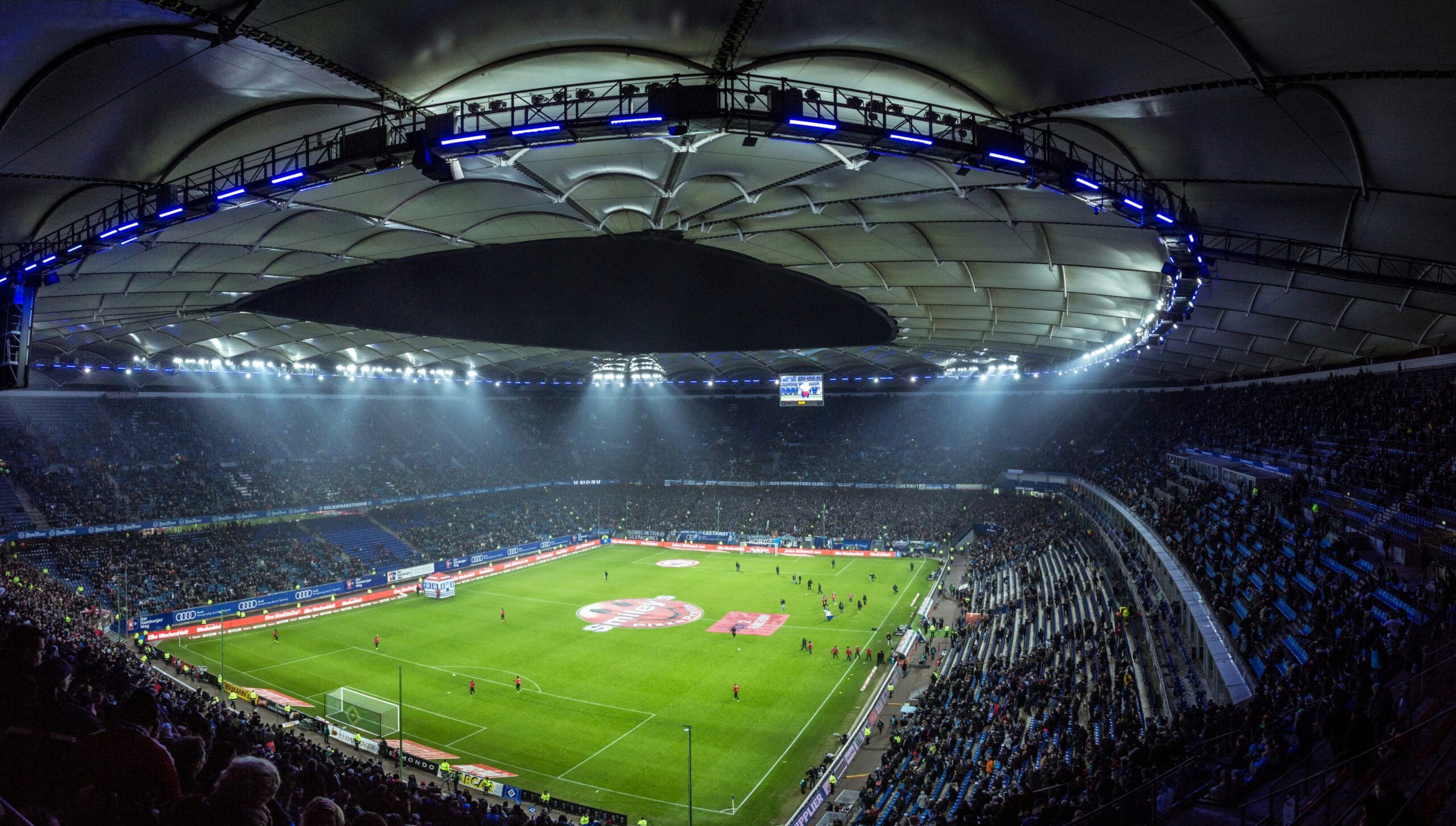 Visão de um estádio lotado da arquibancada, nele está ocorrendo um jogo noturno